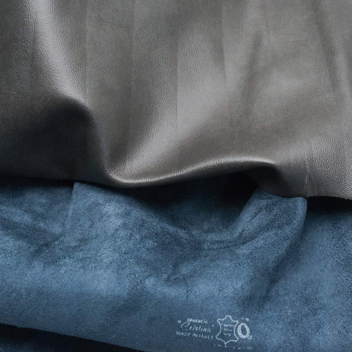 Two folded fabric or leather-like material samples shown close up: a smooth dark gray sample on top and a textured blue sample below. A small white printed logo and text appear on the blue sample near the bottom.