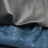 Two folded fabric or leather-like material samples shown close up: a smooth dark gray sample on top and a textured blue sample below. A small white printed logo and text appear on the blue sample near the bottom.