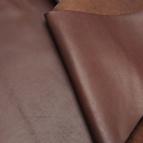 A close-up of a brown leather material, showing its smooth texture and natural grain with a folded corner revealing a slightly warmer tone underneath.